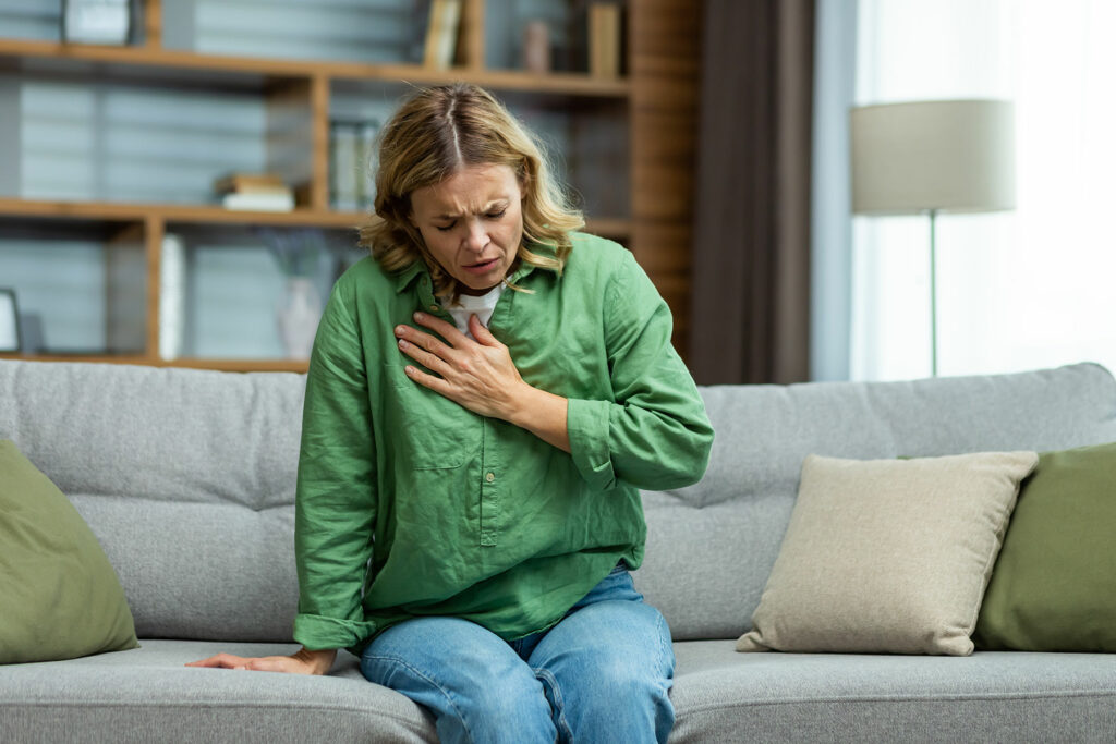 woman sitting on couch holds chest while experiencing the physical signs of anxiety