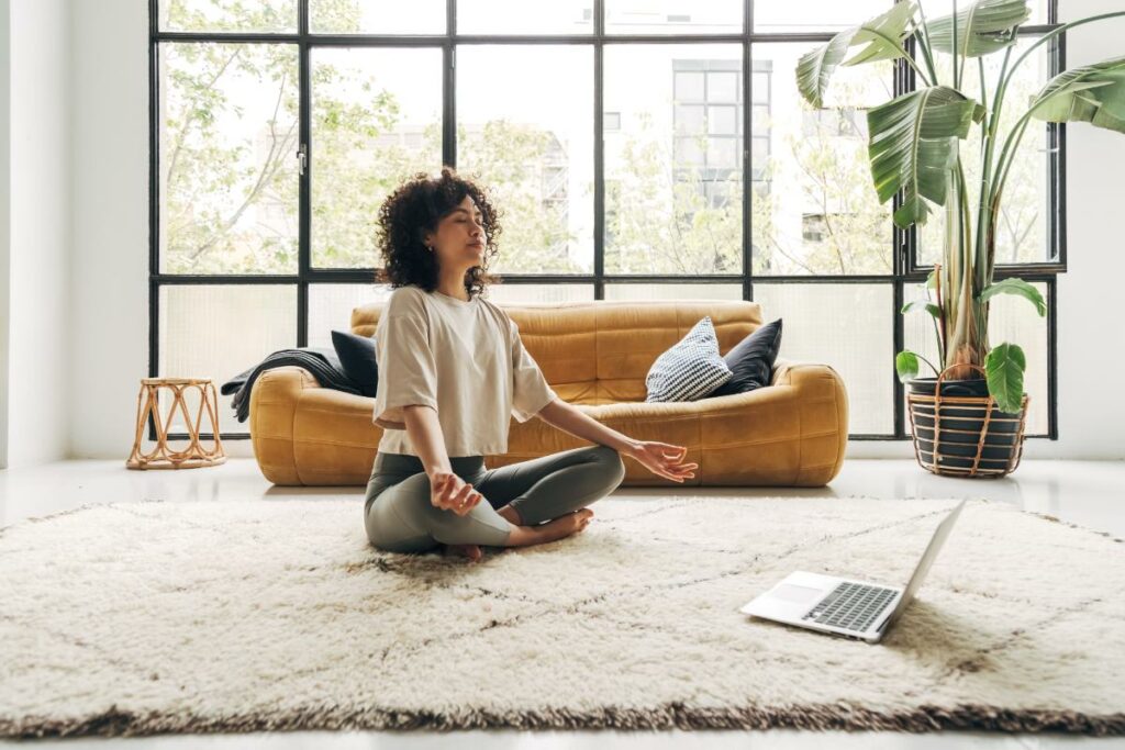 a person meditates in the middle of a room with big windows and a couch to show the benefits of meditation for mental health